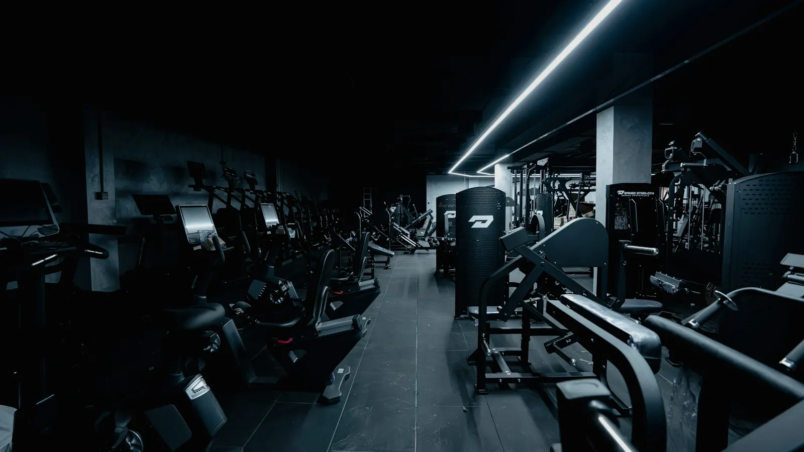 Sala de gimnasio en penumbra con filas de máquinas de cardio y de fuerza